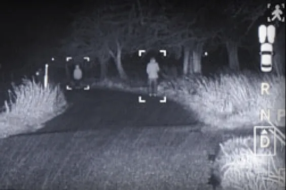 A black and white night-vision view of a road with people standing on the shoulder.